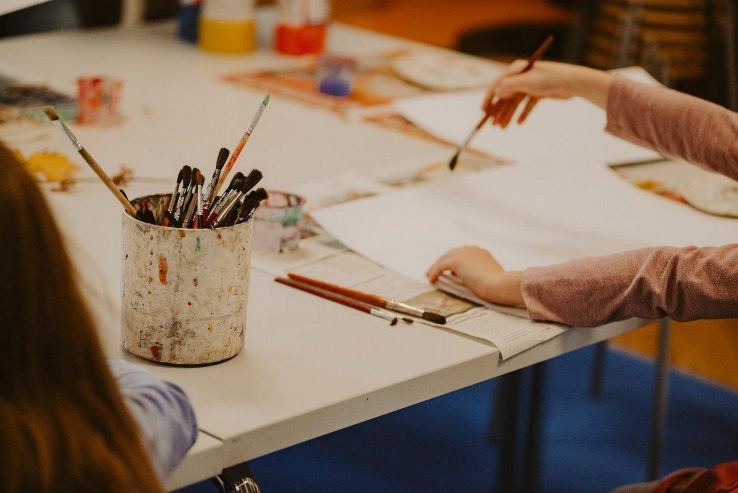 Tisch mit Pinseln und Wasserfarben, zwei Hände eines Kindes sind zu sehen, die mit Pinsel auf Papier malen