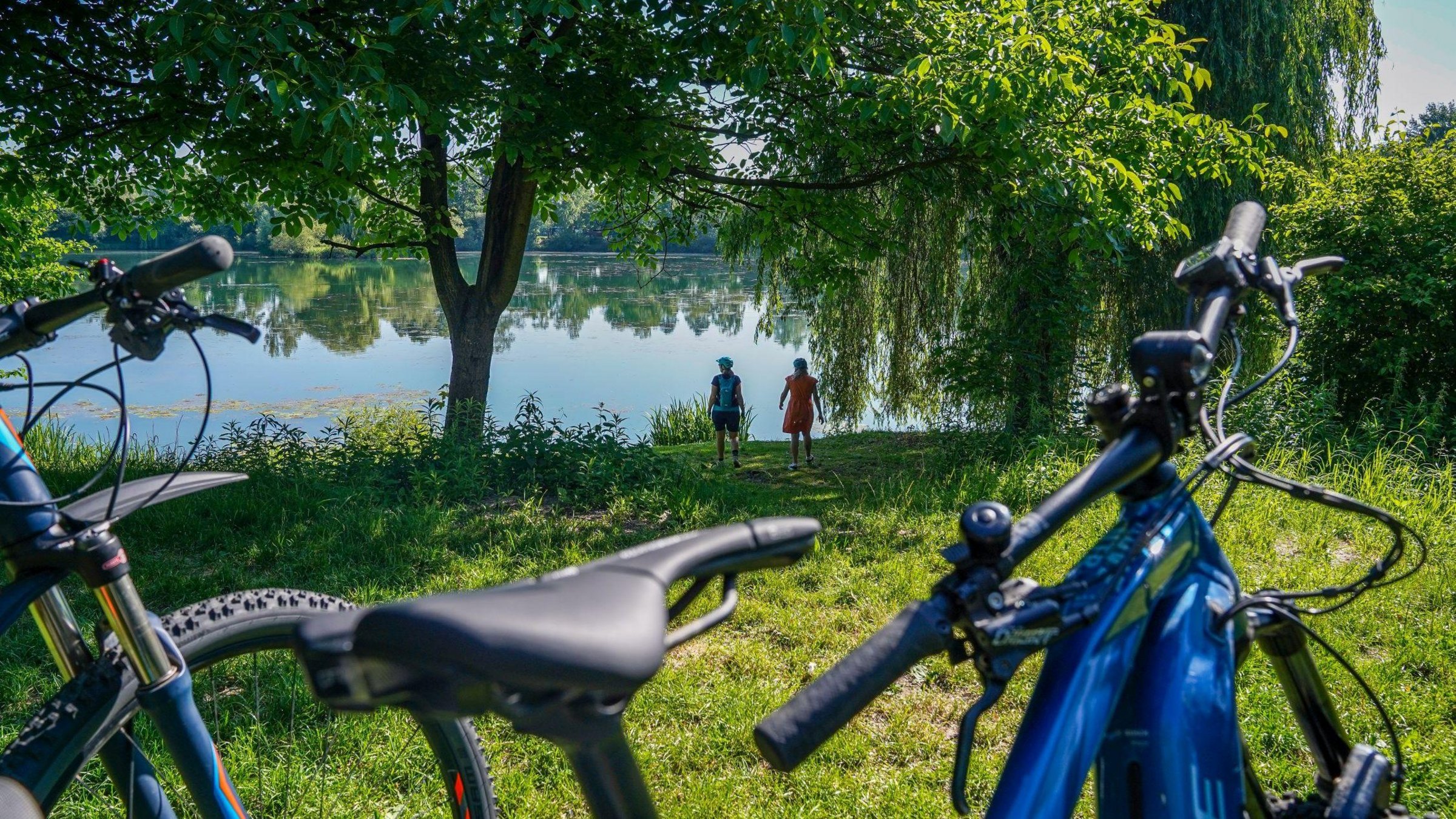 Zwei Fahrräder sind im Vordergrund zu sehen. Im Hintergrund stehen die beiden Fahrer am Ufer eines Sees.