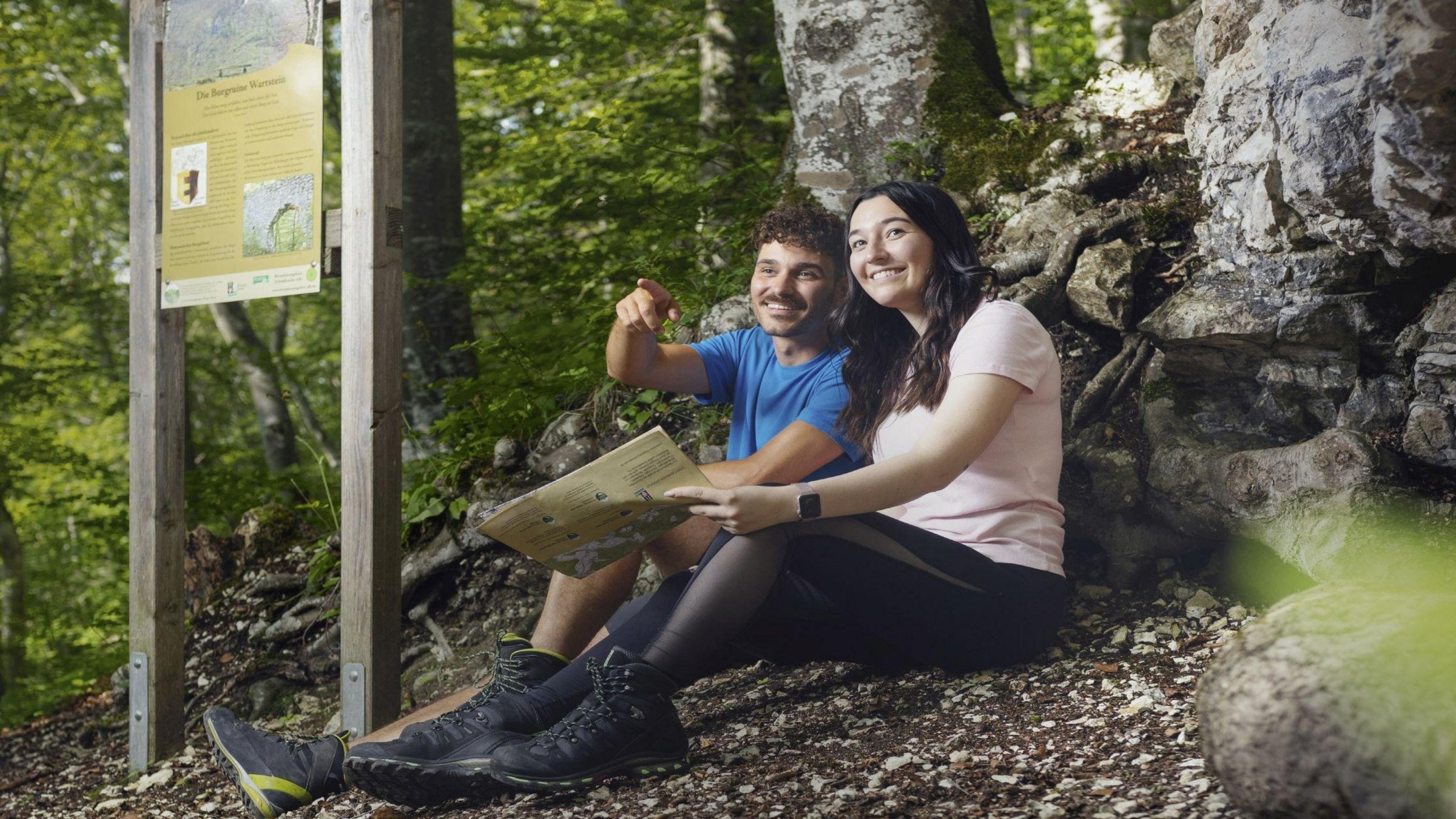 Zwei Personen sitzen an einem Waldweg auf den Felsen. Im Hintergrund ist eine Infotafel zu sehen.